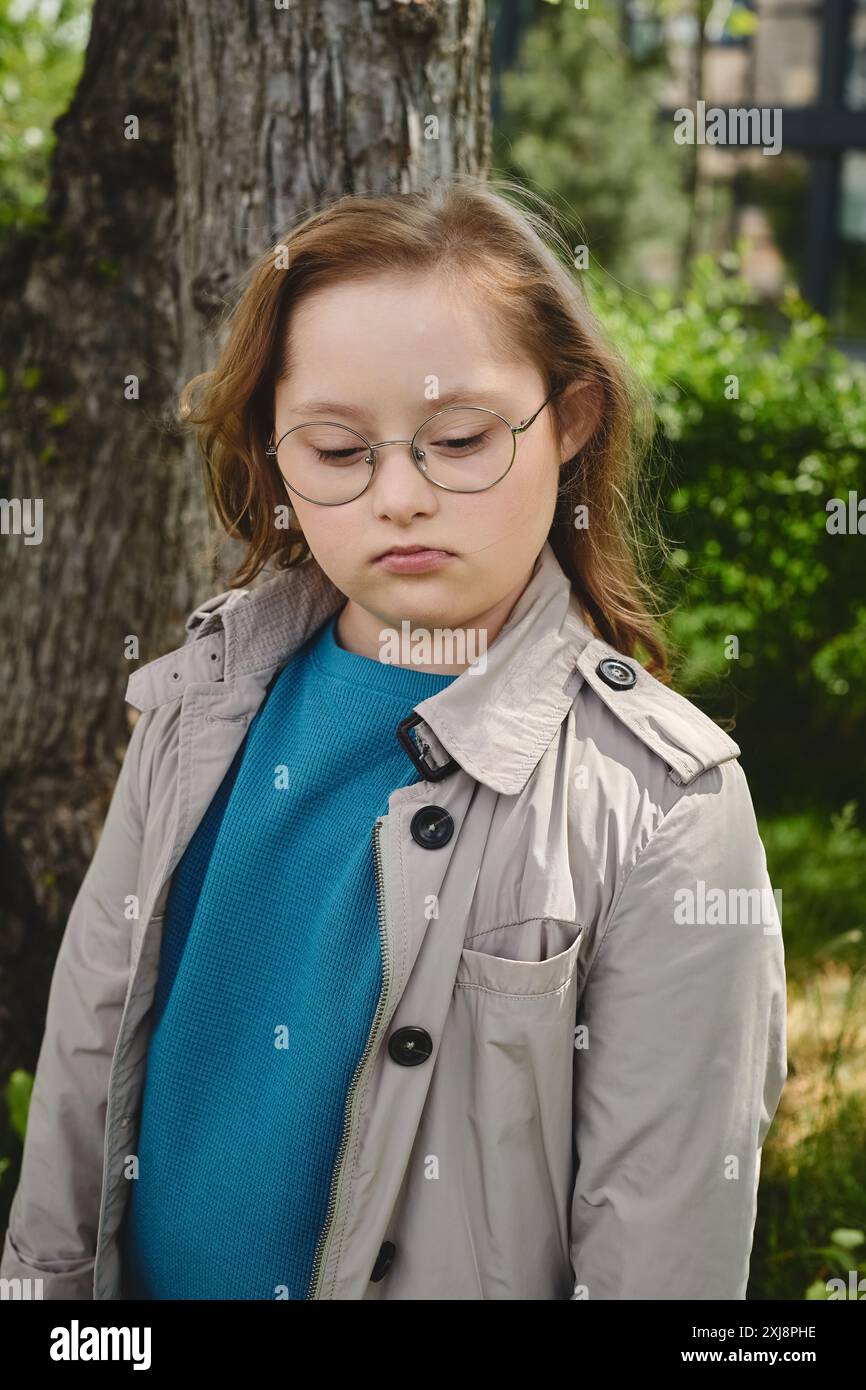 Une petite fille atteinte du syndrome de Down se tient près d'un arbre, perdue dans ses pensées. Banque D'Images