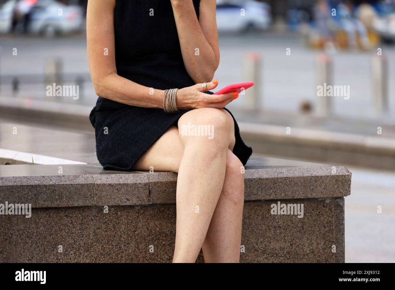 Femme aux jambes croisées portant une robe d'été courte assise avec smartphone sur le banc de rue а Banque D'Images
