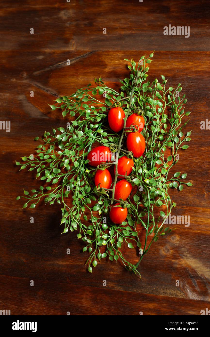 Nature morte de branche allongée de tomates rouges parmi de petites feuilles vertes sur une table en bois brun. Vertical. Banque D'Images