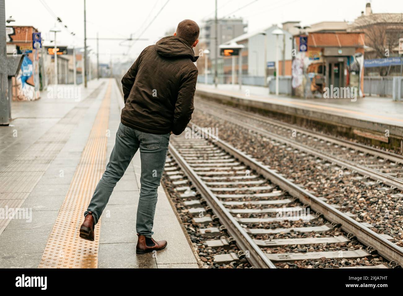 Un homme en jeans et une veste marron se tient sur une plate-forme de train, regardant le long des voies. Banque D'Images