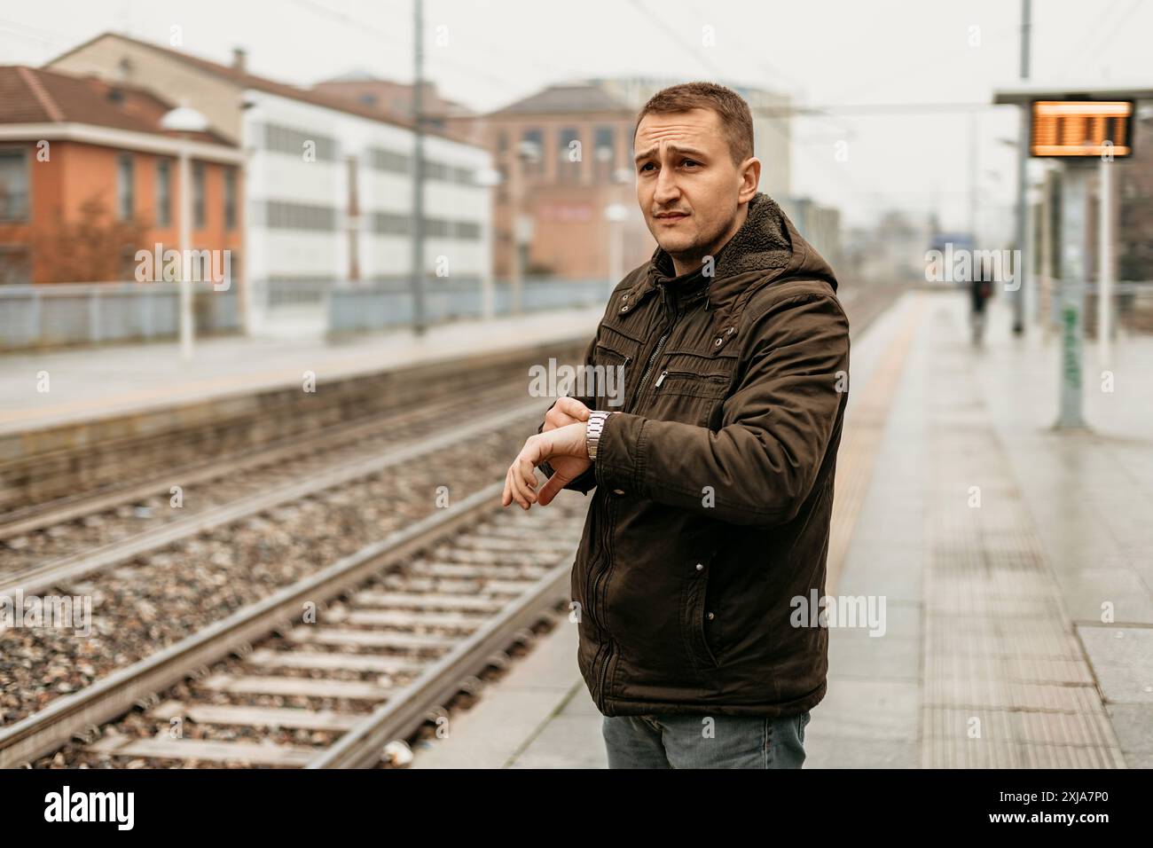 Un homme dans une veste marron vérifie sa montre tout en se tenant debout sur le quai d'une gare. Banque D'Images