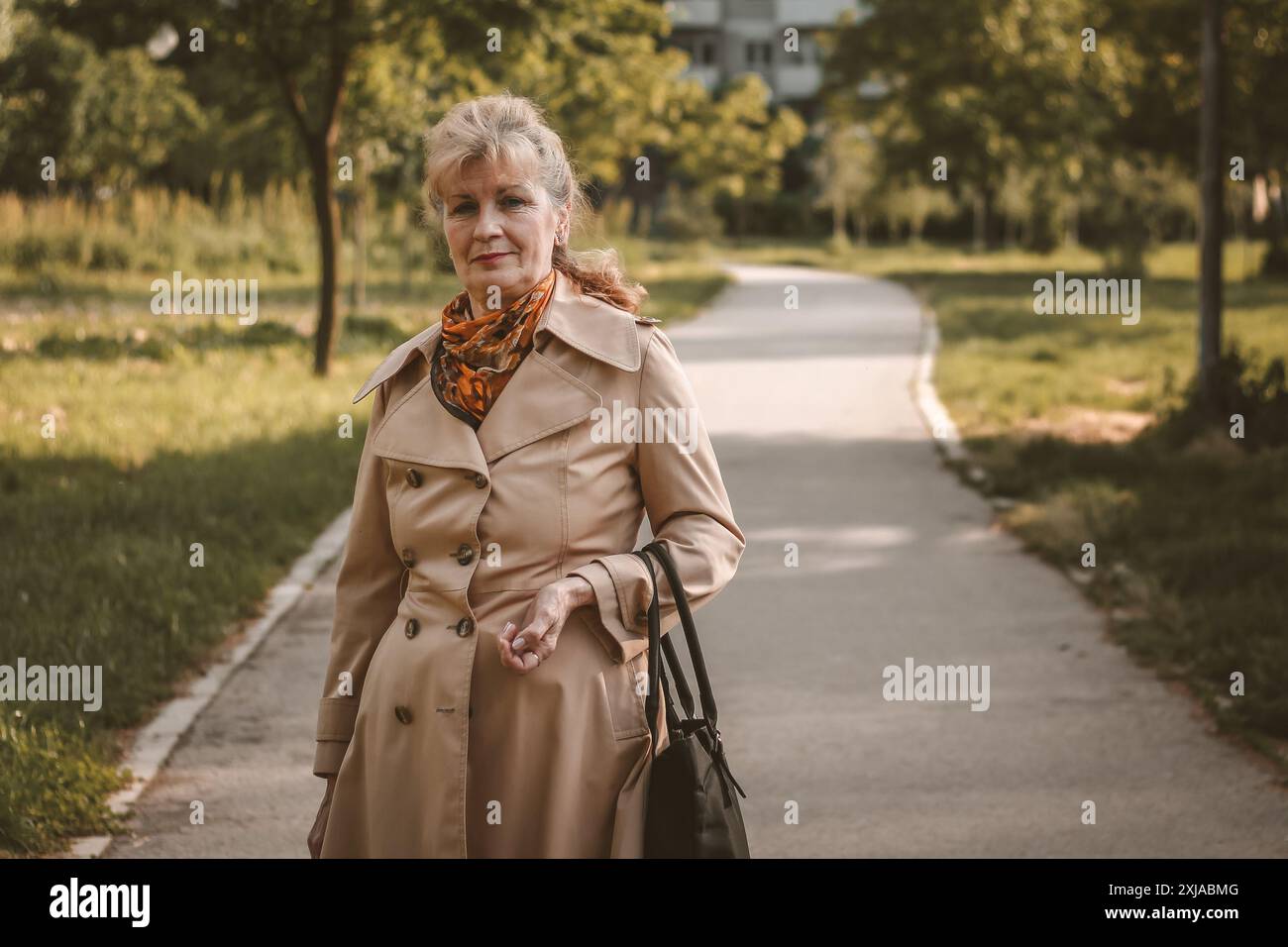 Une femme mature marche le long d'un chemin pavé dans un parc, portant un sac à main noir. Elle porte un trench tan et une écharpe orange. Banque D'Images