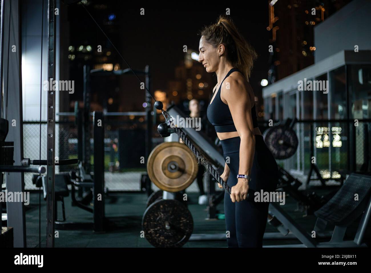 Une femme dans des vêtements d'entraînement noirs effectue un exercice de rangée de câbles dans une salle de gym extérieure la nuit. Banque D'Images