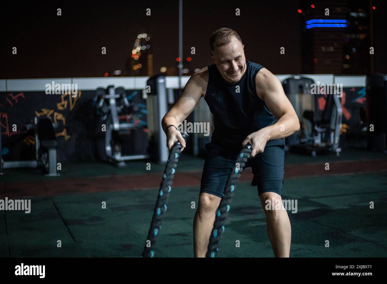 Un homme dans un débardeur noir et des exercices de short avec des cordes de combat sur un gymnase sur le toit la nuit. Banque D'Images