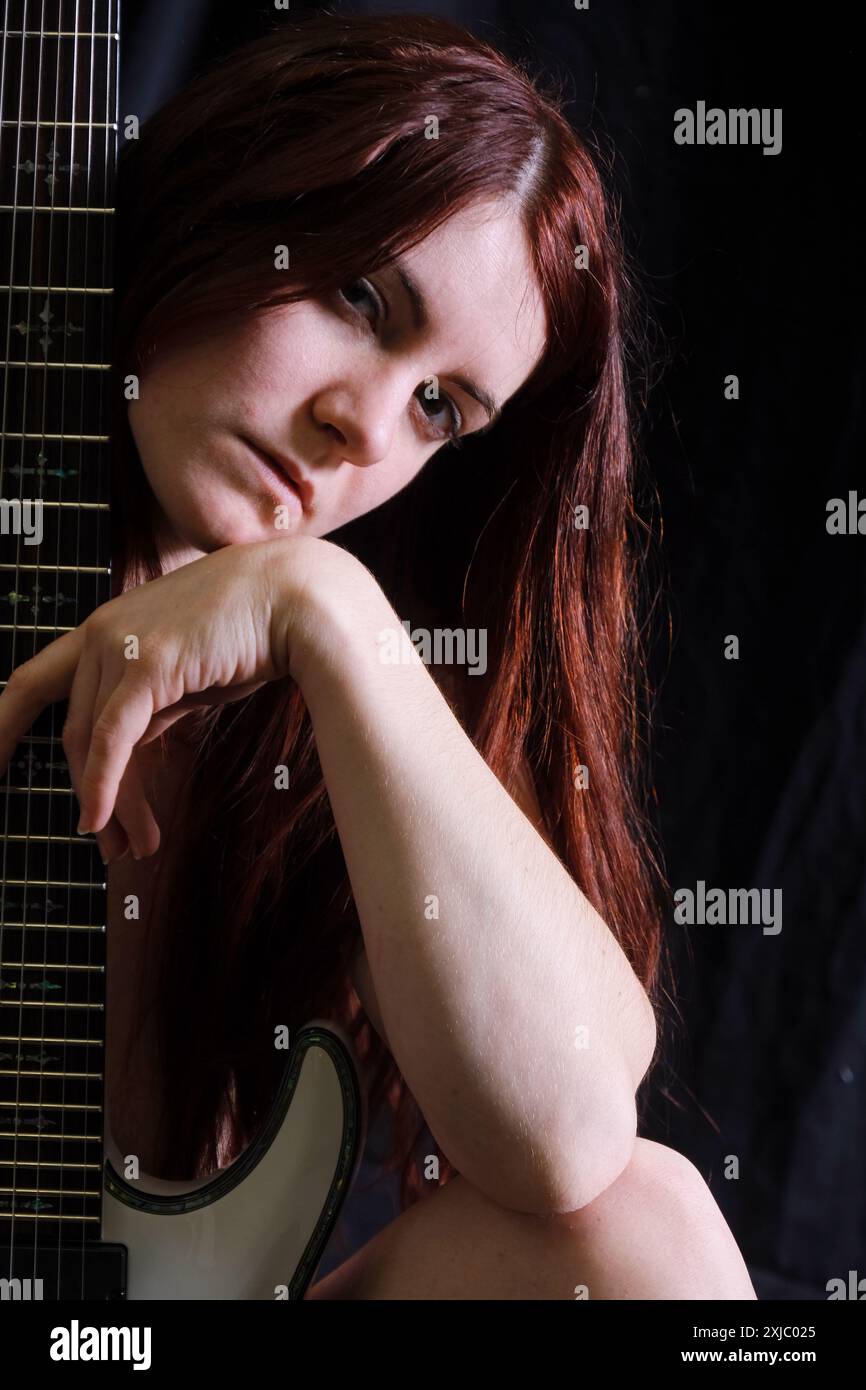 Une femme aux cheveux roux repose la tête et la main sur une guitare électrique, son expression sereine et contemplative. Le fond sombre contraste avec son p Banque D'Images