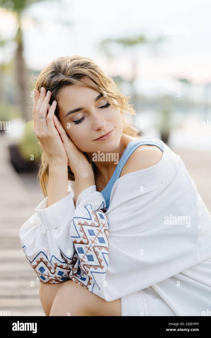 Une femme dans un haut blanc brodé est assise avec sa tête reposant sur ses bras et ses yeux fermés. Banque D'Images