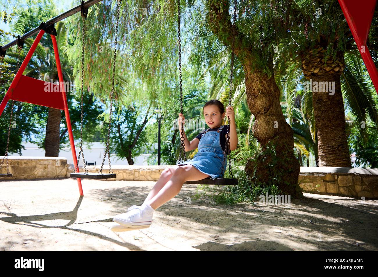 Une jeune fille est assise sur une balançoire dans un parc par une journée ensoleillée, entourée d'arbres et de verdure. Elle semble contente et détendue, profitant de l'activité extérieure Banque D'Images