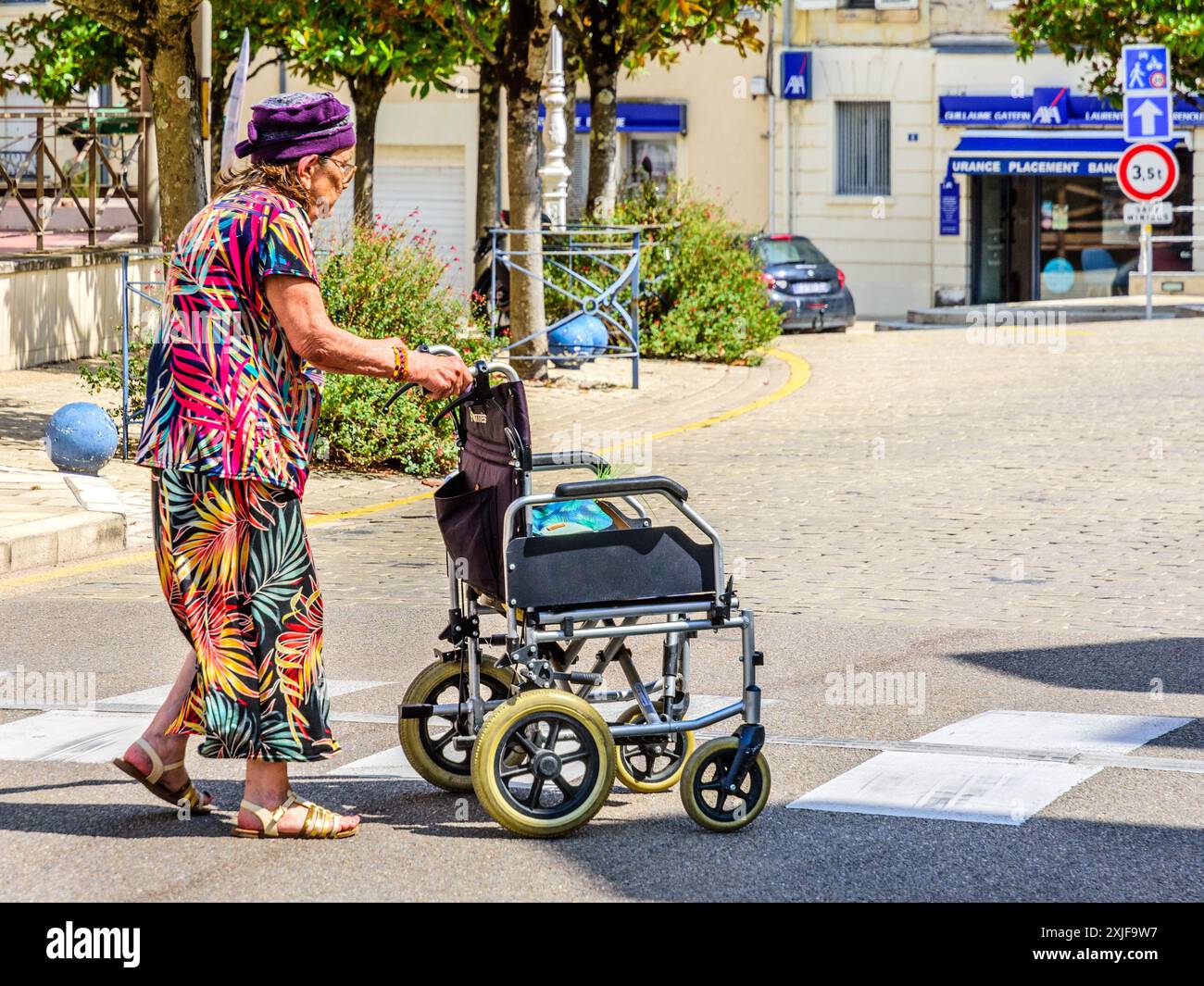 Femme âgée en jupe et haut colorés, poussant son fauteuil roulant à travers la route - la Roche Posay, Vienne (86), France. Banque D'Images