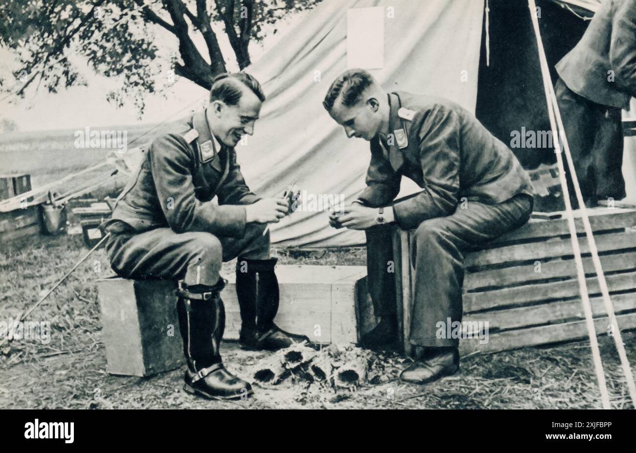 Une photographie montre des soldats allemands dans un camp temporaire, assis à l'extérieur de leur tente et préparant des crêpes de pommes de terre (Kartoffelpuffer). Cette scène offre un aperçu de leur vie quotidienne lors de l’opération Barbarossa en 1941, alors qu’ils avançaient vers le front de l’est. Banque D'Images