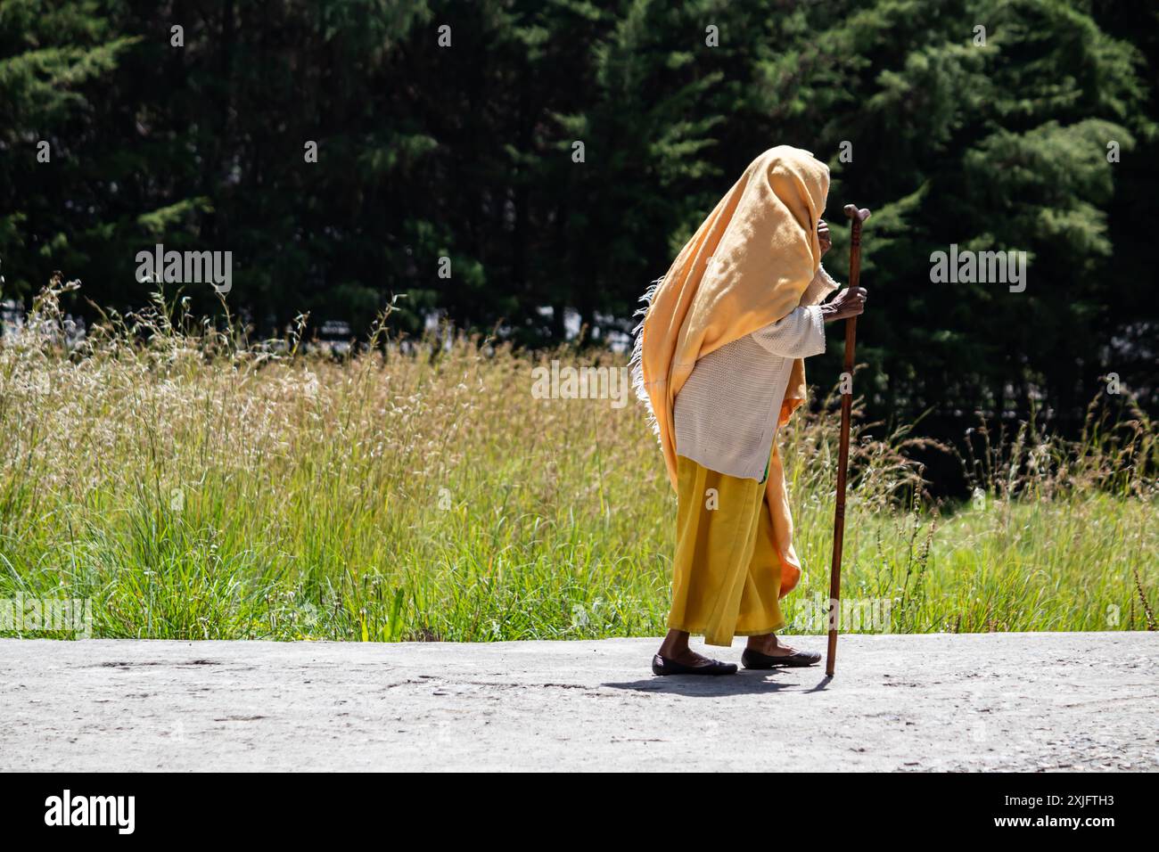 Une vieille maman éthiopienne, traditionnellement vêtue de jaune, marche avec une canne en bois, reflétant le patrimoine culturel, la dignité et la résilience Banque D'Images