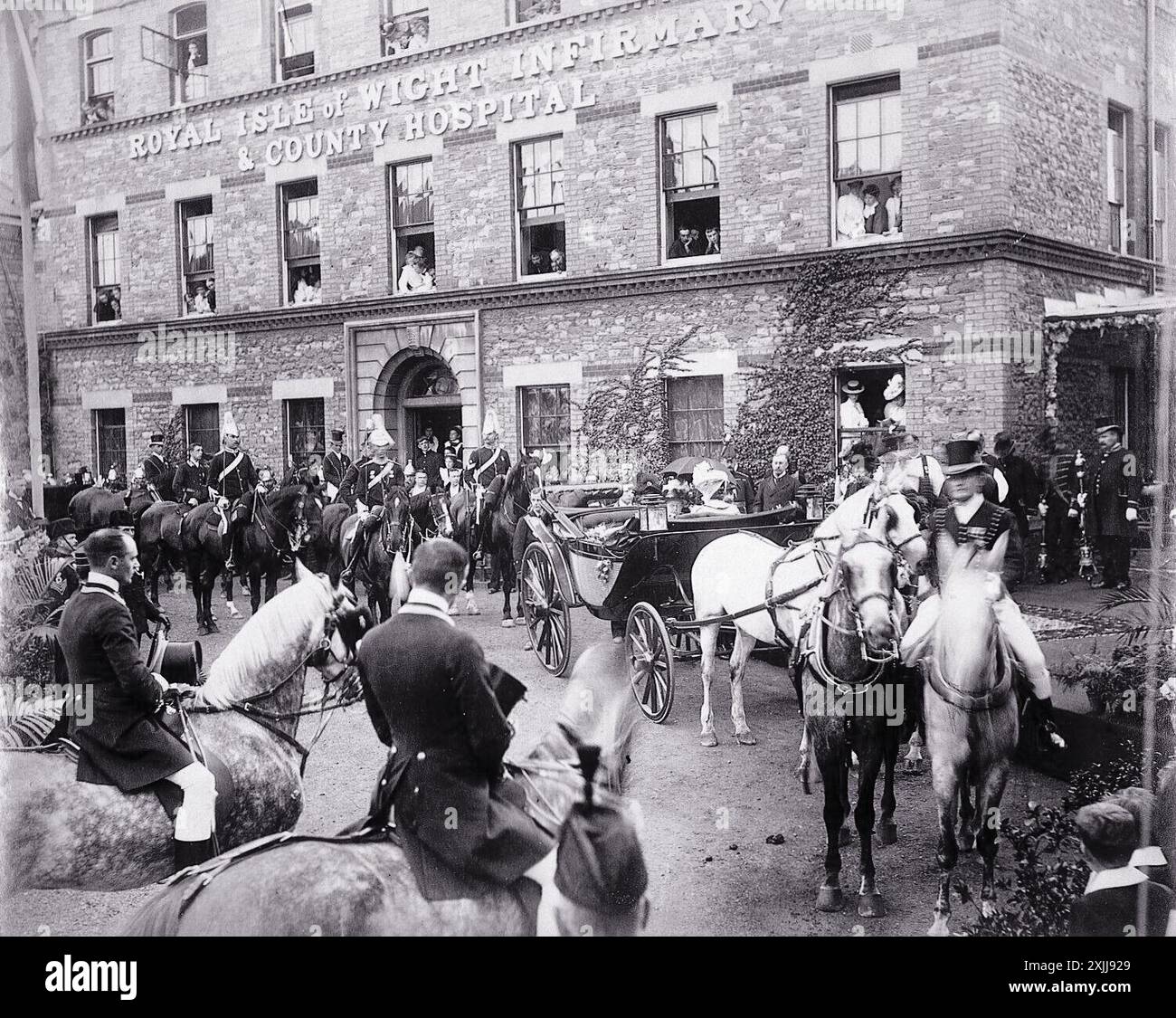 La reine Victoria, accompagnée de préposés, arrive à l'infirmerie royale de l'île de Wight et à l'hôpital du comté en 1899. La photographie montre la reine dans une calèche tirée par des chevaux, avec un groupe de cavaliers qui l'accompagne. Le bâtiment de l'hôpital est visible en arrière-plan, les gens regardant par les fenêtres. Banque D'Images