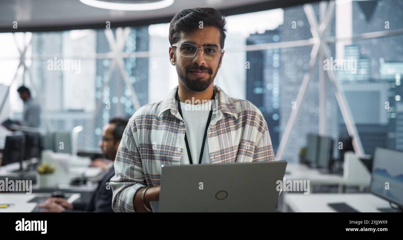 Bel homme indien portant des lunettes, souriant charmante et regardant la caméra. Jeune ingénieur intelligent tenant un ordinateur portable. Directeur financier travaillant dans le siège social moderne Banque D'Images