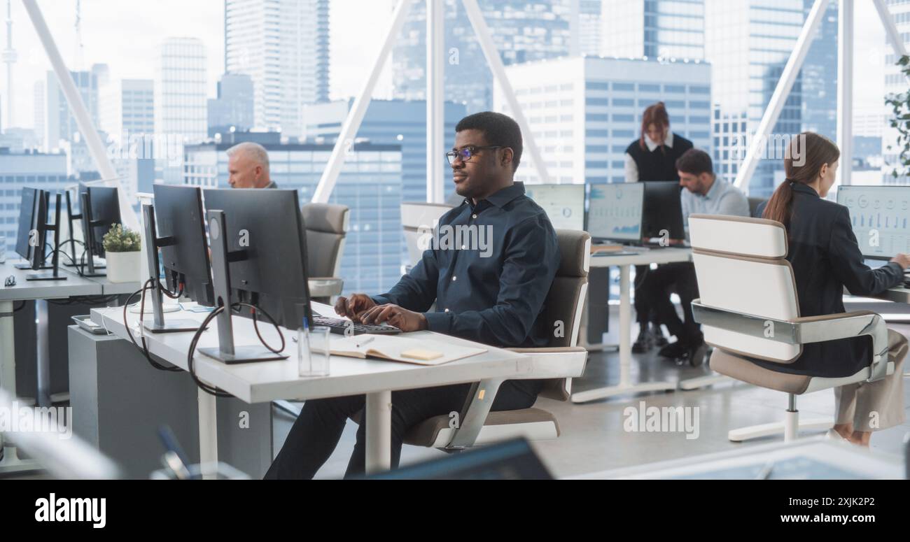 Analyste de données masculin afro-américain ciblé tapant sur ordinateur de bureau dans divers bureaux d'entreprise avec Megapolis Window View. Professionnel Balck homme travaillant dans la société de conseil avec des collègues. Banque D'Images