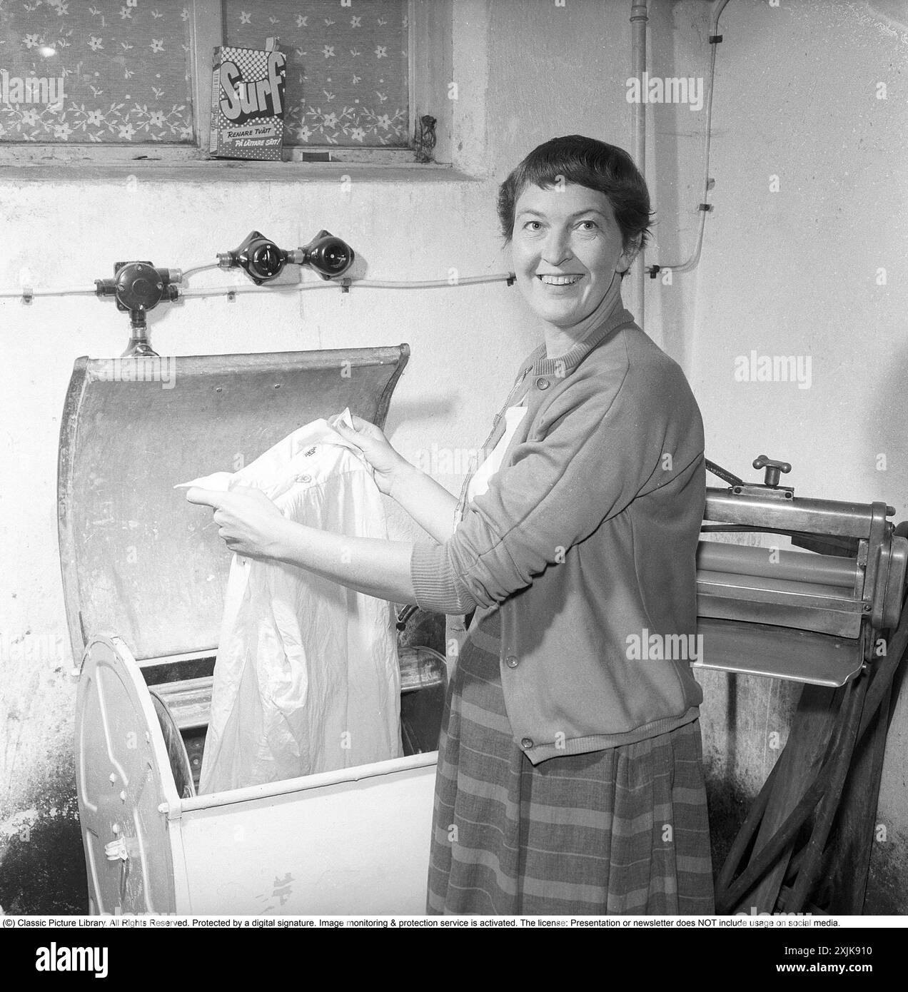 Dans la buanderie 1955. Une femme est vue dans la buanderie avec un vêtement dans la machine à laver. À cette époque dans les années 1950, la fonction spin n'existait pas. L'eau dans les vêtements a été mutilée, cette mangle peut être vu sur la droite. Un paquet de détergent est dans la fenêtre du sous-sol. Surfez avec le texte : plus de blanchisserie d'une manière plus facile! Un détergent lancé sur le marché suédois dans les années 1950 Anders Svahn ref SVA12 Banque D'Images