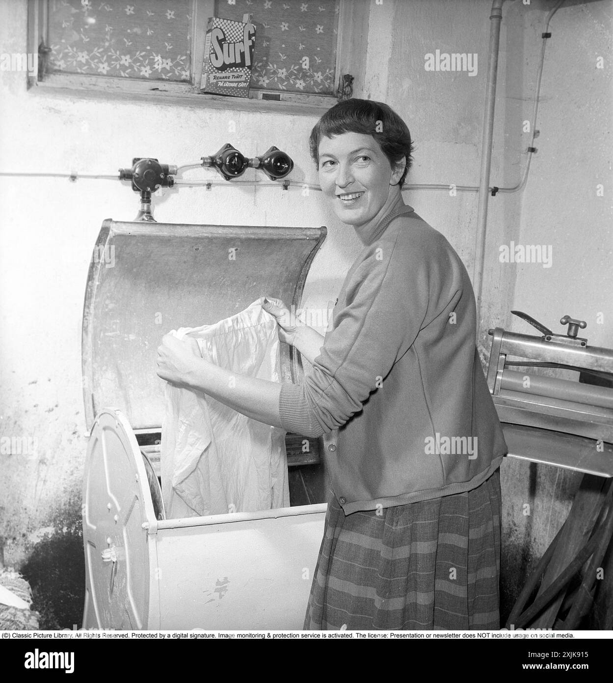 Dans la buanderie 1955. Une femme est vue dans la buanderie avec un vêtement dans la machine à laver. À cette époque dans les années 1950, la fonction spin n'existait pas. L'eau dans les vêtements a été mutilée, cette mangle peut être vu sur la droite. Un paquet de détergent est dans la fenêtre du sous-sol. Surfez avec le texte : plus de blanchisserie d'une manière plus facile! Un détergent lancé sur le marché suédois dans les années 1950 Anders Svahn ref SVA12 Banque D'Images