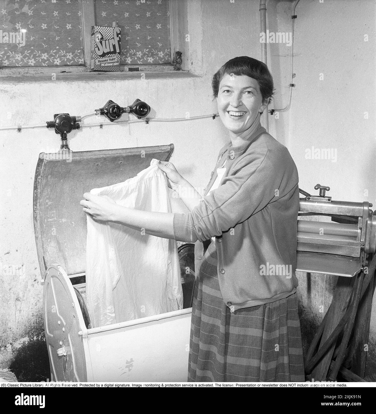 Dans la buanderie 1955. Une femme est vue dans la buanderie avec un vêtement dans la machine à laver. À cette époque dans les années 1950, la fonction spin n'existait pas. L'eau dans les vêtements a été mutilée, cette mangle peut être vu sur la droite. Un paquet de détergent est dans la fenêtre du sous-sol. Surfez avec le texte : plus de blanchisserie d'une manière plus facile! Un détergent lancé sur le marché suédois dans les années 1950 Anders Svahn ref SVA12 Banque D'Images