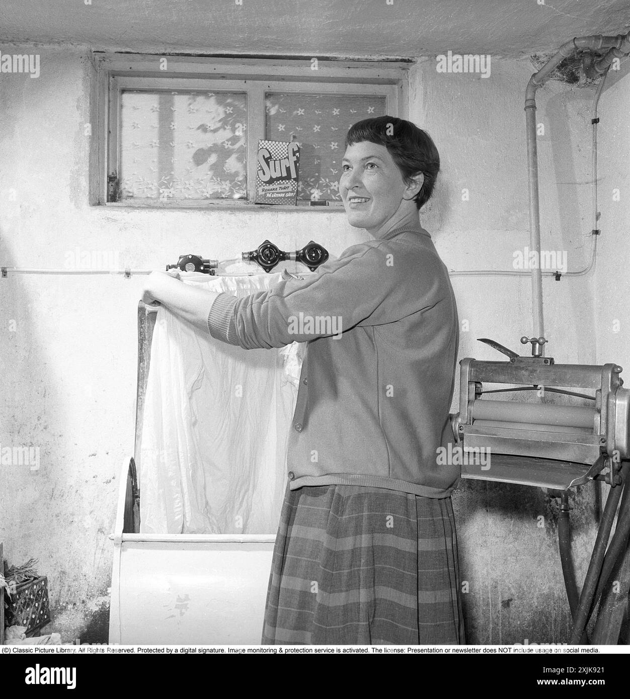 Dans la buanderie 1955. Une femme est vue dans la buanderie avec un vêtement dans la machine à laver. À cette époque dans les années 1950, la fonction spin n'existait pas. L'eau dans les vêtements a été mutilée, cette mangle peut être vu sur la droite. Un paquet de détergent est dans la fenêtre du sous-sol. Surfez avec le texte : plus de blanchisserie d'une manière plus facile! Un détergent lancé sur le marché suédois dans les années 1950 Anders Svahn ref SVA12 Banque D'Images