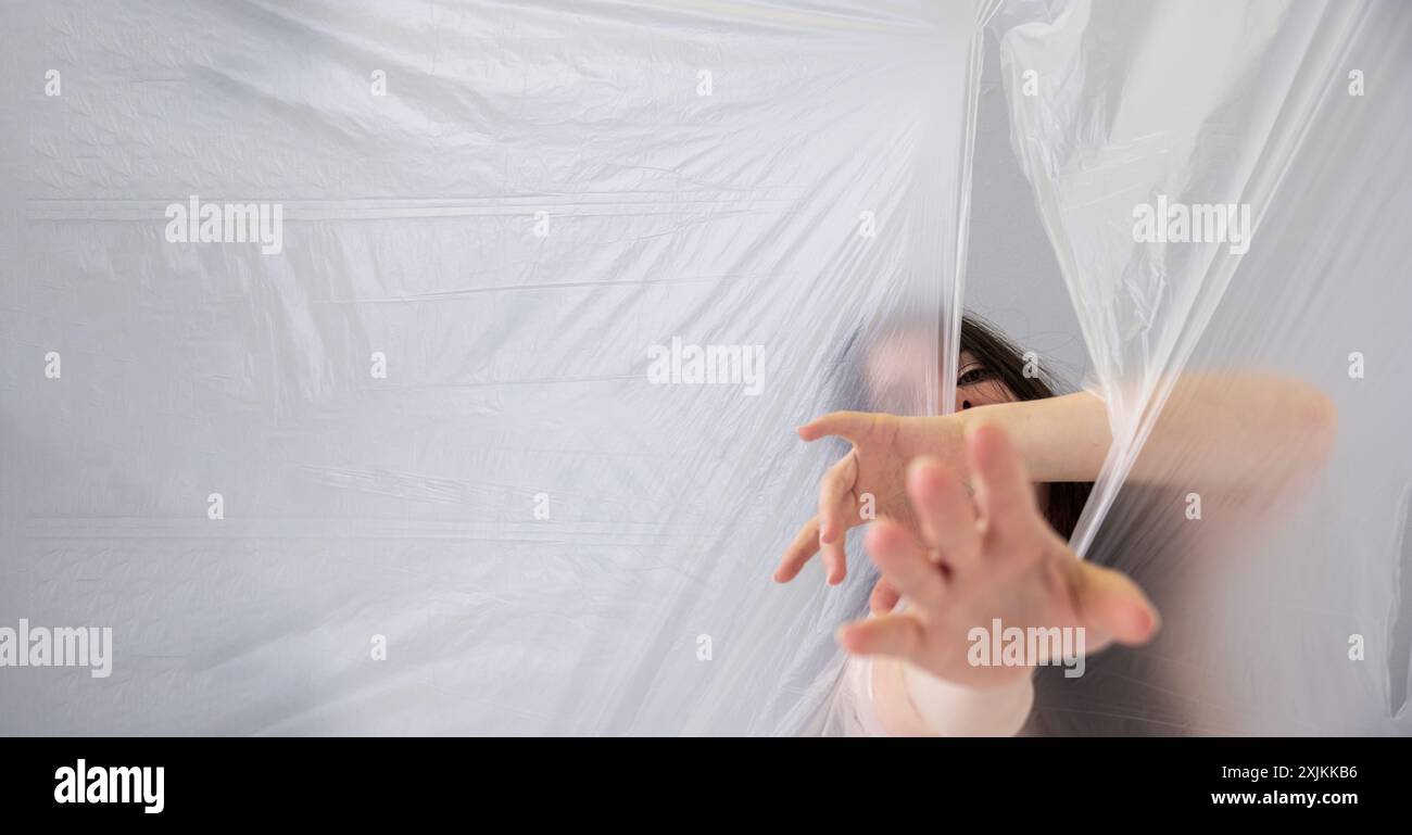 Une femme tend la main par derrière une feuille de plastique translucide, créant un sentiment de mystère et de lutte. Sa main tendue et partiellement obscurcie fa Banque D'Images