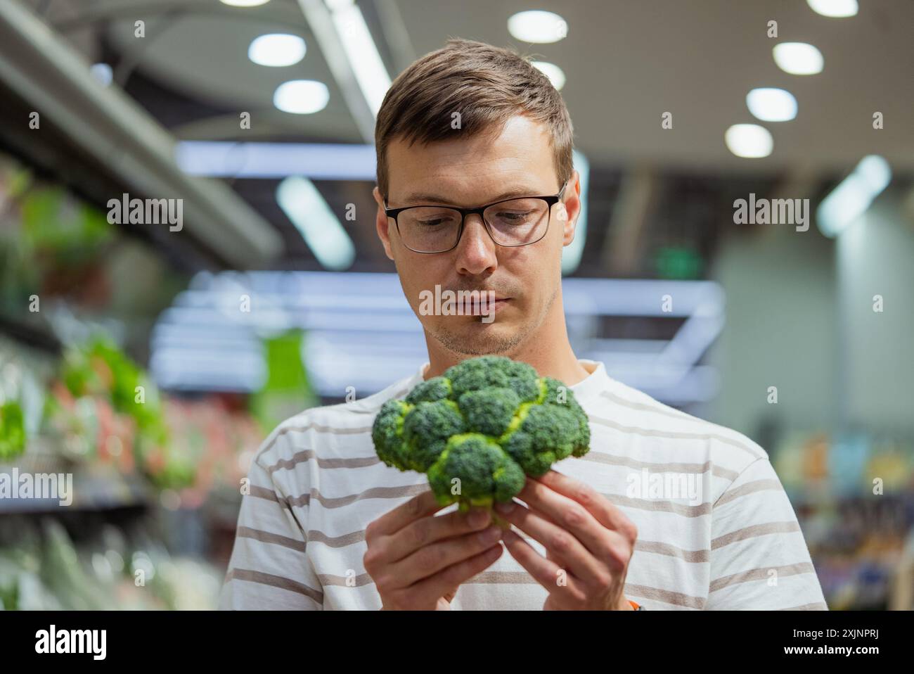 Homme en chemise rayée et lunettes inspectant une tête de brocoli fraîche tout en faisant des courses dans la section des produits d'un supermarché. Banque D'Images