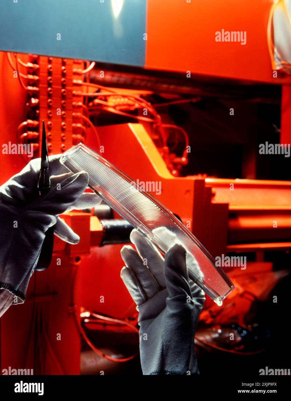 Un travailleur portant des gants blancs tient une pièce de voiture en plastique transparent devant une machine de fabrication rouge. La pièce semble être un feu arrière ou un pavillon Banque D'Images