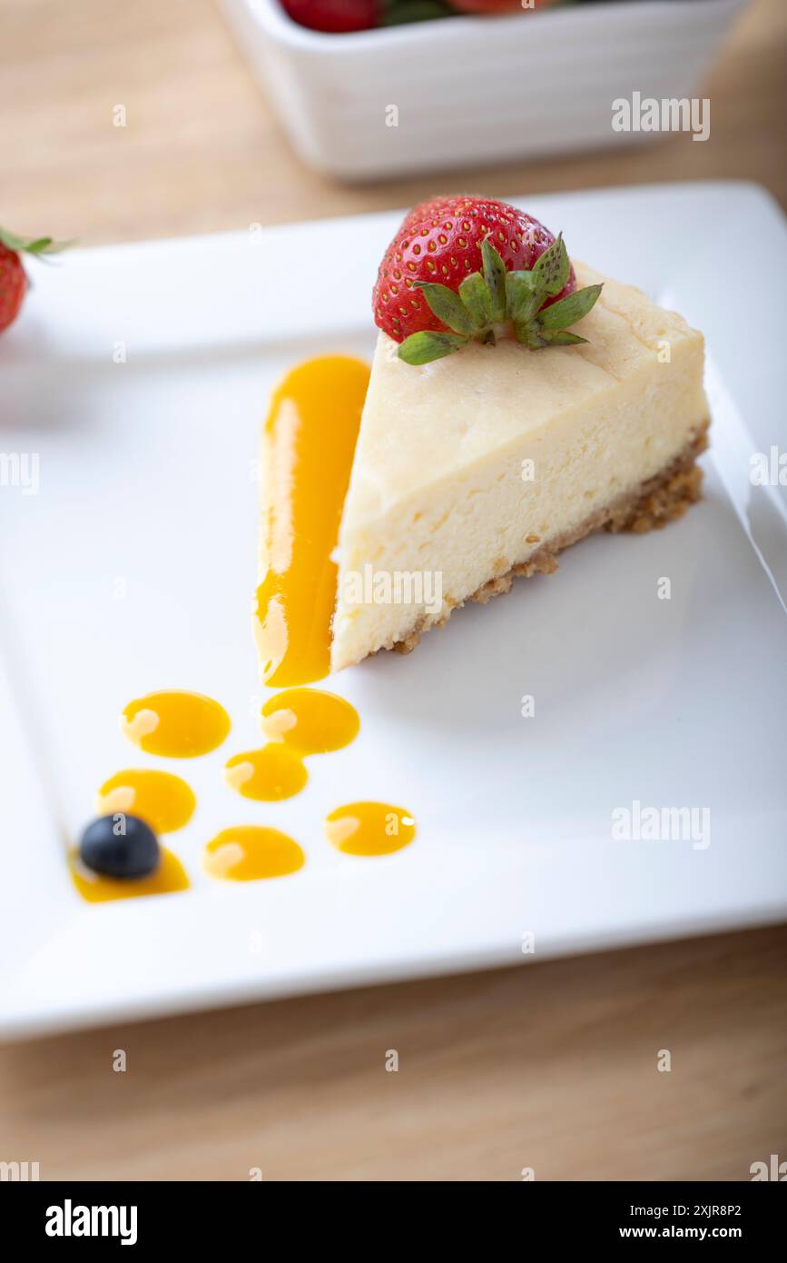 Une tranche de cheesecake crémeux garnie d’une fraise fraîche, servie sur une assiette blanche avec un filet de sauce aux fruits. Un bol de fraises fraîches Banque D'Images