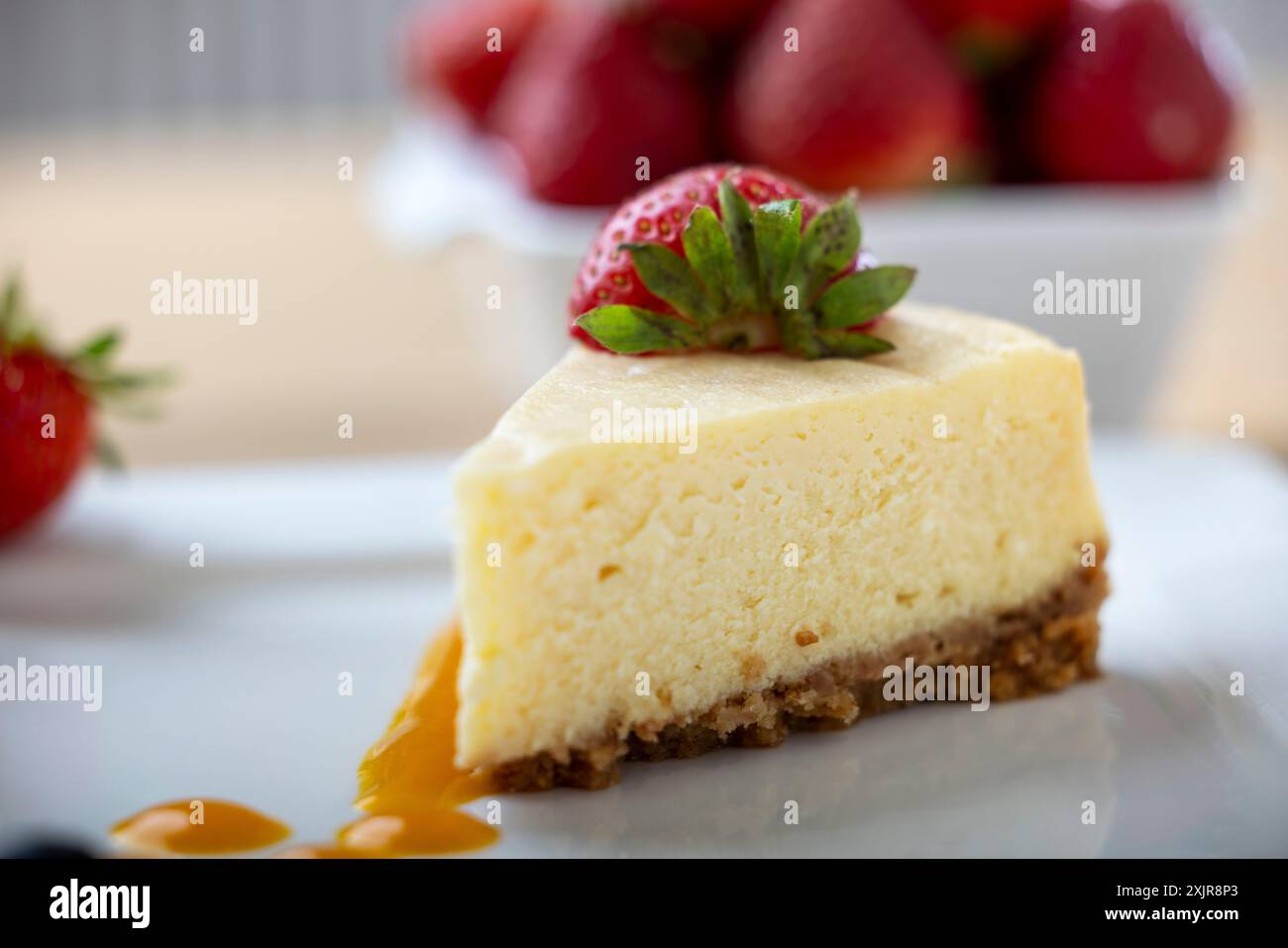 Une tranche de cheesecake crémeux garnie d’une fraise fraîche, servie sur une assiette blanche avec un filet de sauce aux fruits. Un bol de fraises fraîches Banque D'Images