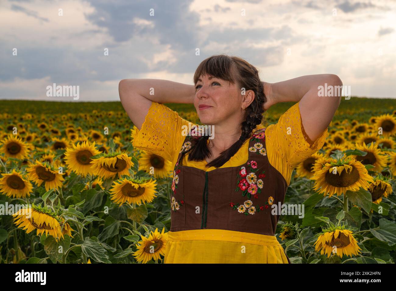 Une femme se tient dans un champ avec des tournesols jaunes. Ses cheveux sont tressés, elle porte une blouse jaune, une robe de soleil brune et un tablier jaune. Banque D'Images