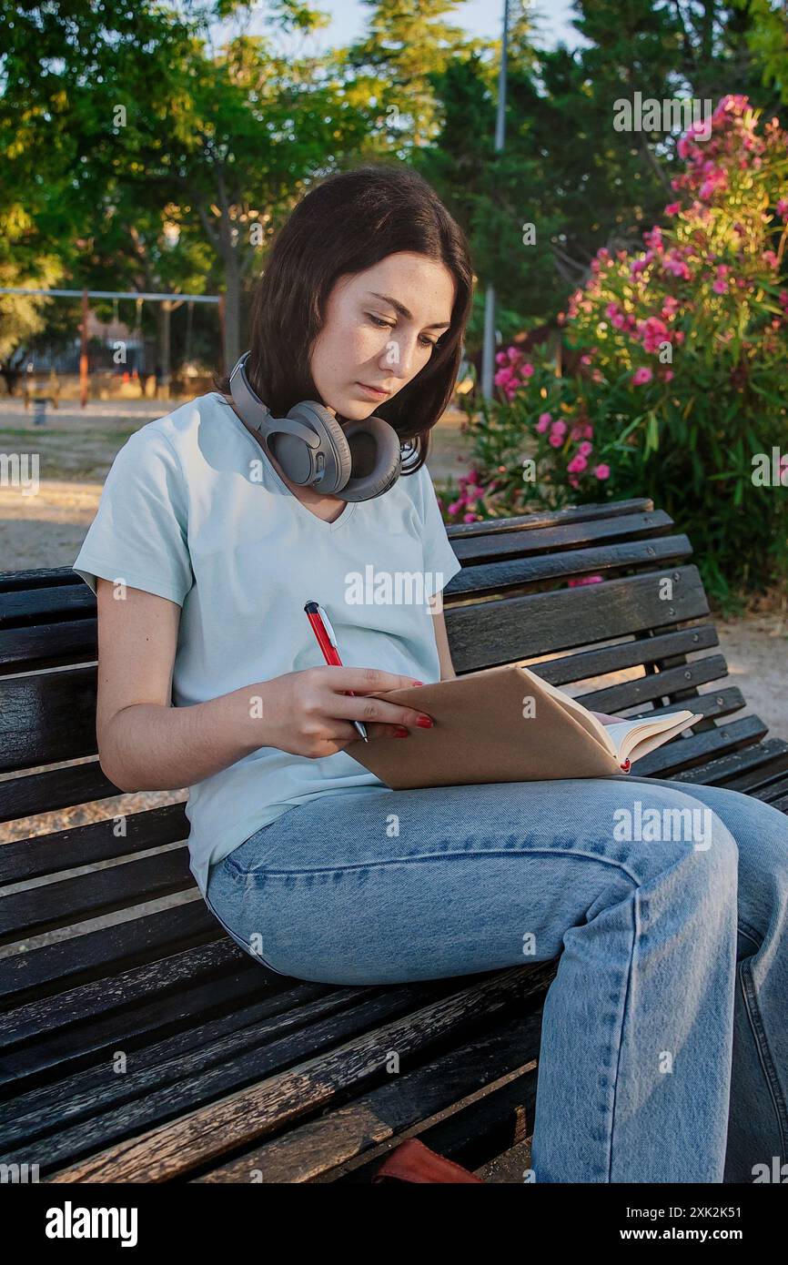Une jeune femme se concentre sur l'écriture dans son cahier alors qu'elle est assise sur un banc de parc, avec des écouteurs autour du cou et un fond serein de rose f Banque D'Images