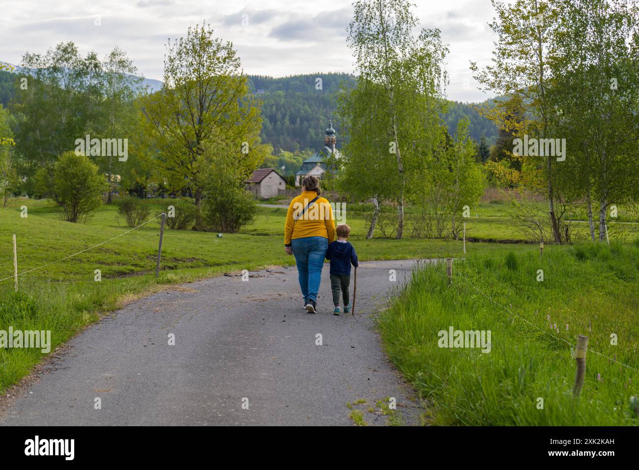 Un enfant heureux tient la main de sa mère alors qu'ils marchent main dans la main le long d'un chemin rural. Un petit garçon tient un bâton dans ses mains. Parentalité : mère et fils Banque D'Images