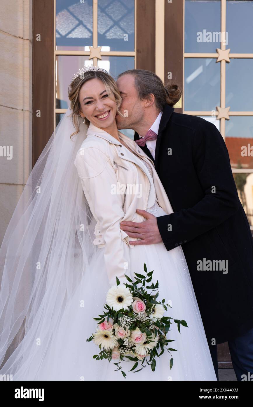 Le marié embrasse doucement la mariée sur le cou, la mariée regarde la caméra et rit joyeusement Алекс целует Banque D'Images