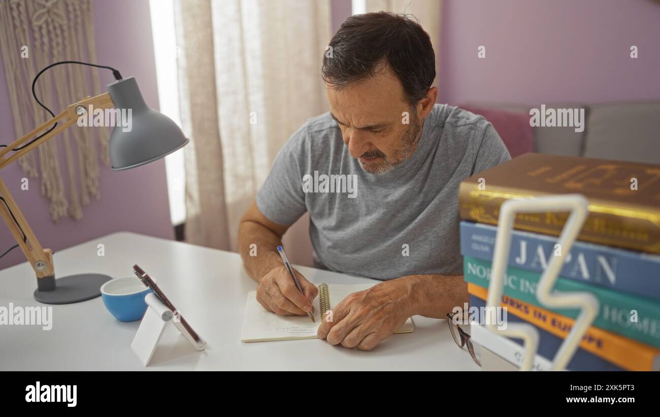 Homme hispanique d'âge moyen écrivant dans un cahier à la maison avec des livres sur un bureau, se concentrant intensément dans un salon très éclairé. Banque D'Images