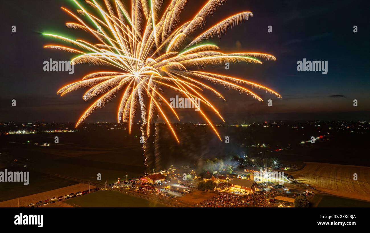 Les feux d'artifice dorés rayonnent brillamment au cours D'Un rassemblement festif, illuminant le ciel nocturne et illuminant le parc d'expositions animé ci-dessous, entouré de vastes champs et de paysages ruraux. Banque D'Images