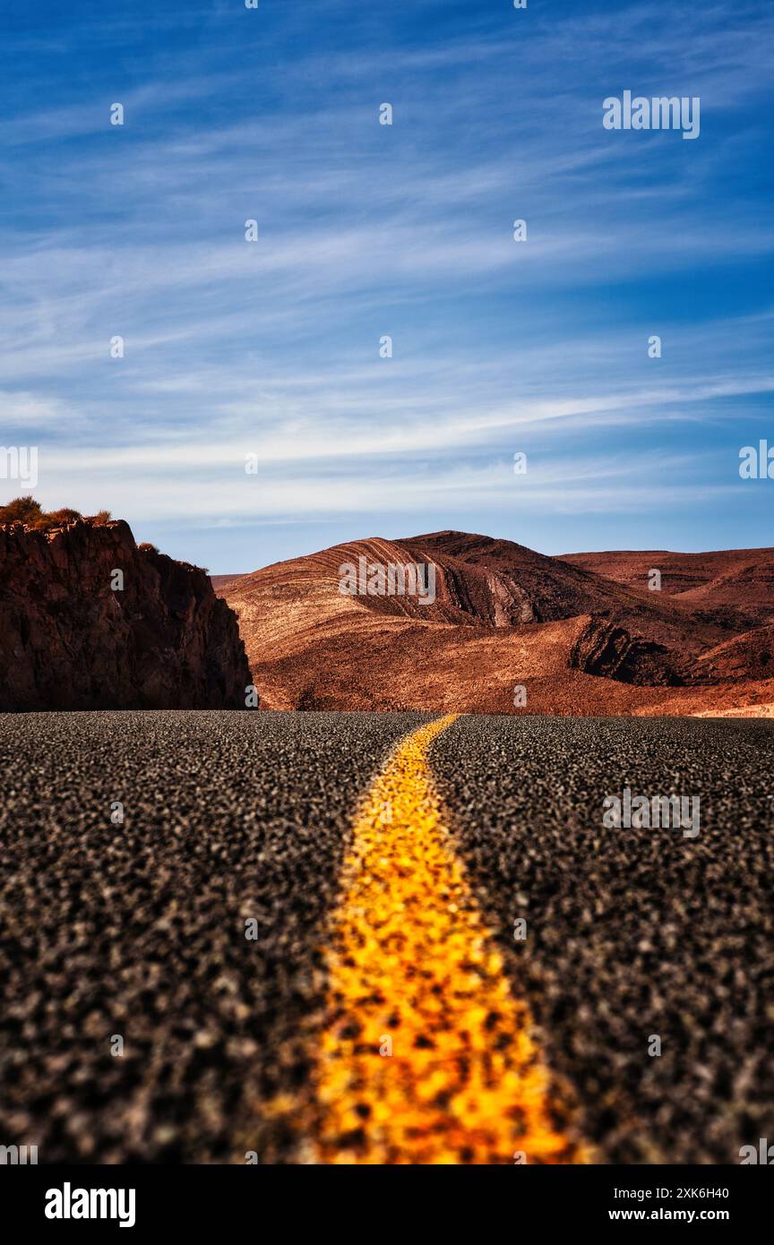 La route ouverte menant à travers le magnifique paysage désertique marocain. Ligne jaune guidant le chemin, les montagnes escarpées et le ciel expansif. Banque D'Images