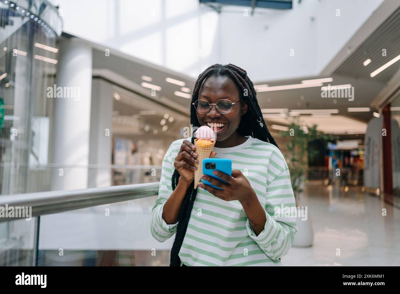 Femme souriante et profitant d'un cône de crème glacée tout en textant sur son smartphone à l'intérieur d'un centre commercial moderne. Black Girl mange un dessert sucré froid en faisant du shopping. Banque D'Images