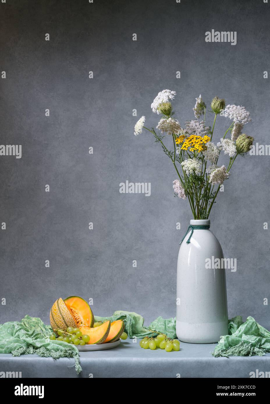 Nature morte avec des fleurs. Fleurs sauvages blanches et jaunes dans un grand vase clair sur un fond gris clair, à côté de lui se trouve un melon coupé et un grand bouquet de W. Banque D'Images