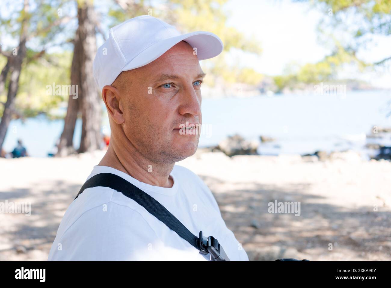 Homme portant une casquette blanche et un t-shirt blanc, regardant au loin. Des arbres et un lac sont en arrière-plan. Concept de loisirs de plein air Banque D'Images