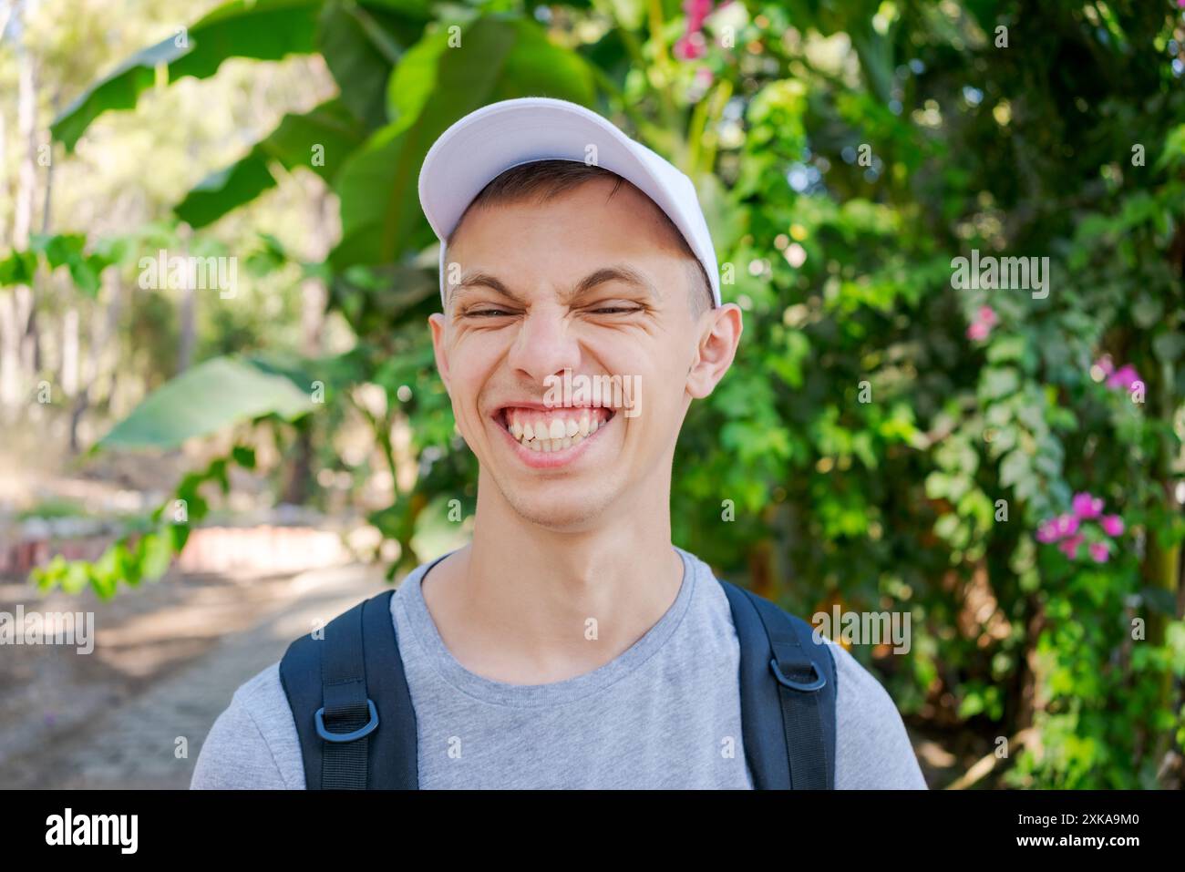 Jeune homme portant une casquette et un sac à dos, souriant les yeux fermés dans un parc verdoyant. Concept de bonheur et de plaisir en plein air Banque D'Images