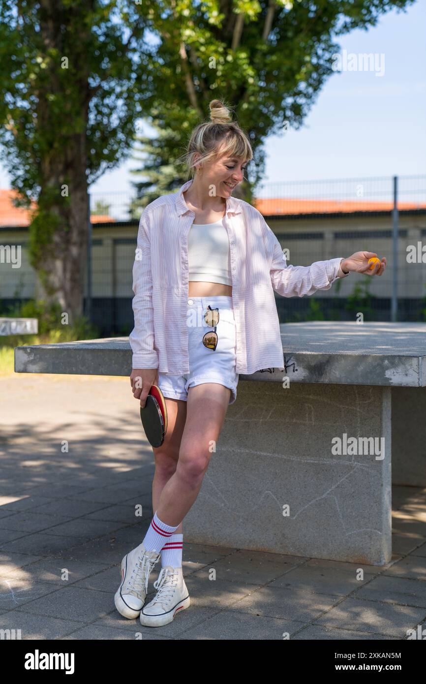 Une fille moderne et souriante en short court est assise sur une table de tennis dans la rue, tient deux raquettes de tennis de table rouges dans ses mains Banque D'Images