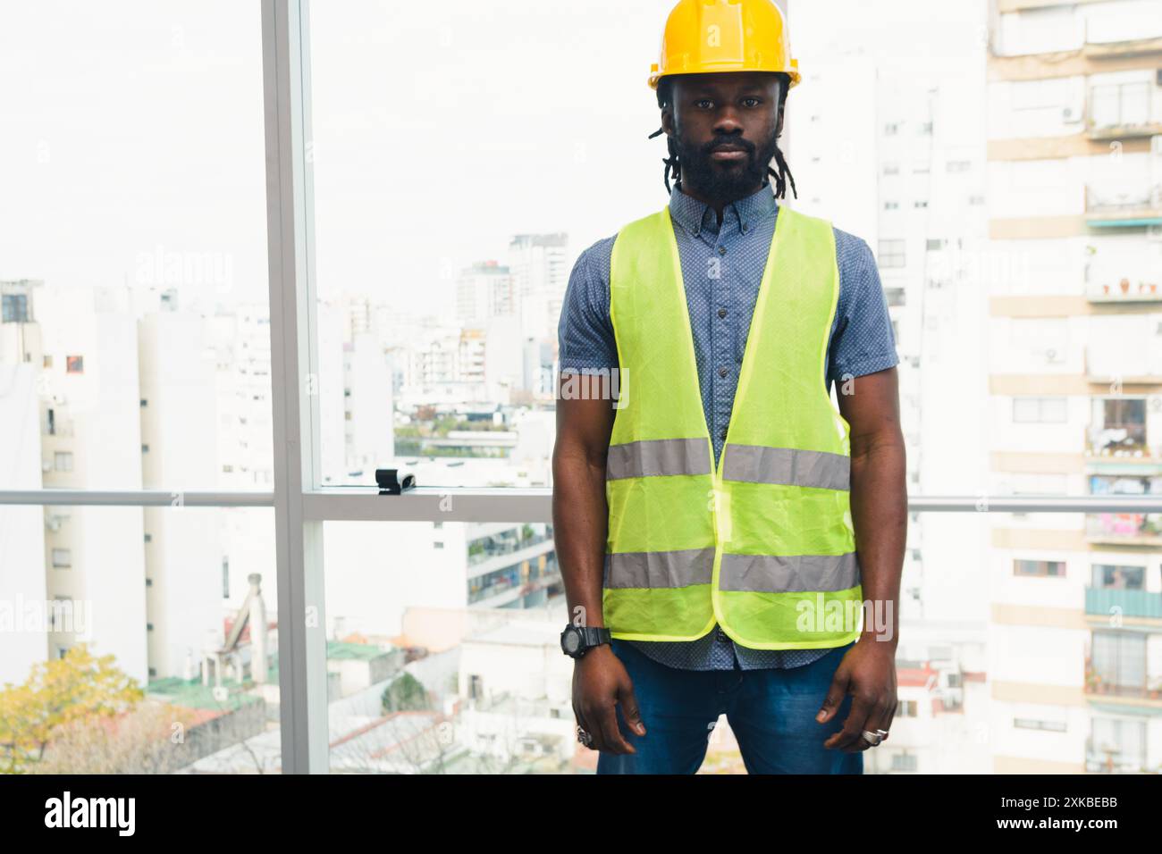Portrait de jeune ingénieur africain debout dans le bureau regardant la caméra, il porte un casque réfléchissant et un casque de sécurité jaune, il a la fenêtre b Banque D'Images
