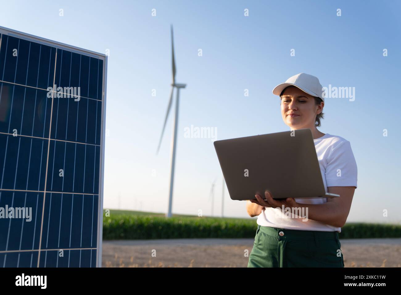 Agriculteur femme portant une casquette blanche et un t-shirt avec des supports pour ordinateur portable à côté du panneau solaire. Éoliennes en arrière-plan. Banque D'Images