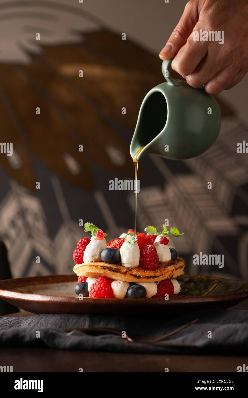Le sirop d'érable est versé sur une pile de crêpes avec des fraises et des baies sur le dessus. La main de l'homme coulant. Banque D'Images