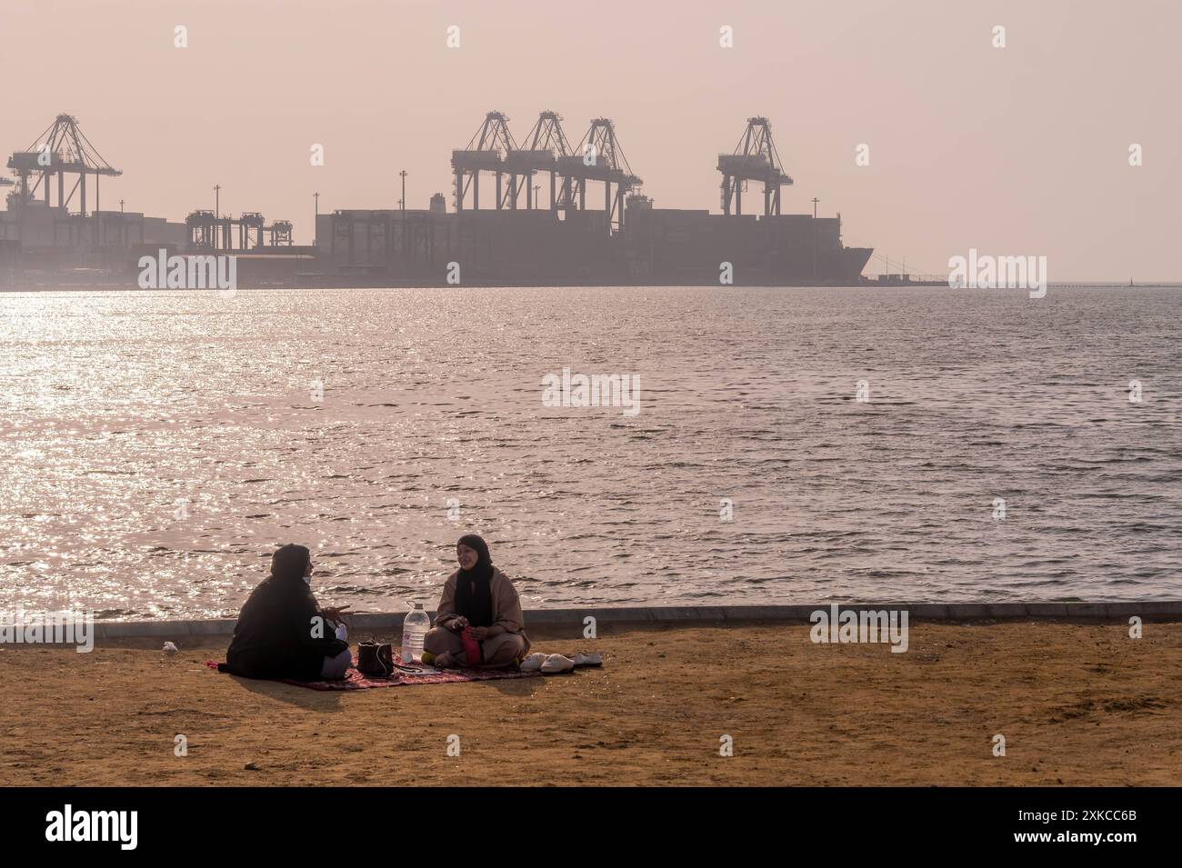 Deux femmes musulmanes assises sur la côte de la mer Rouge avec vue sur le port islamique de Djeddah en Arabie Saoudite avec les grues portuaires et les terminaux de navires. Banque D'Images