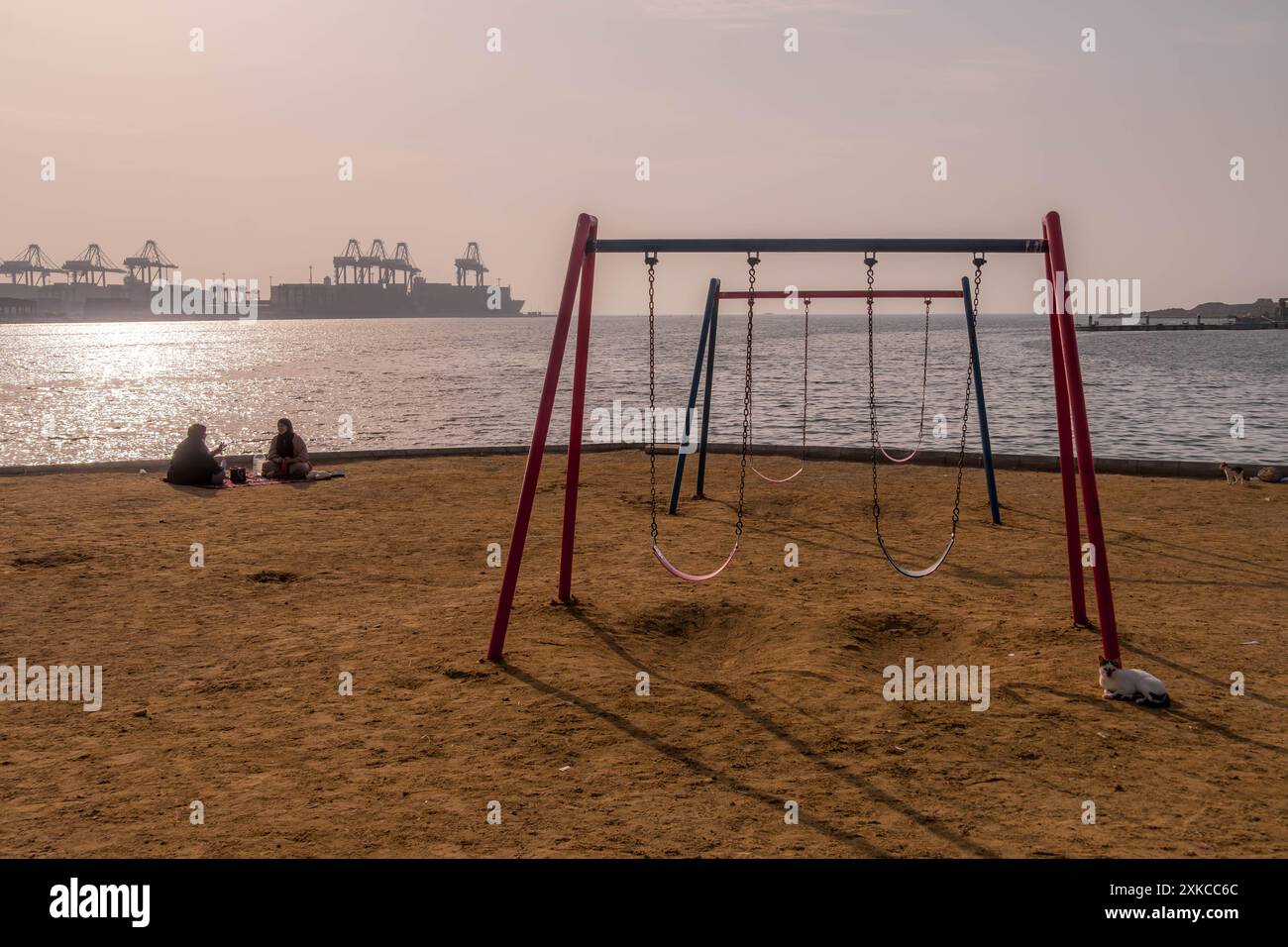Deux femmes musulmanes assises sur une aire de jeux près de la côte de la mer Rouge avec la vue du port islamique de Djeddah en Arabie Saoudite avec les grues du port maritime. Banque D'Images