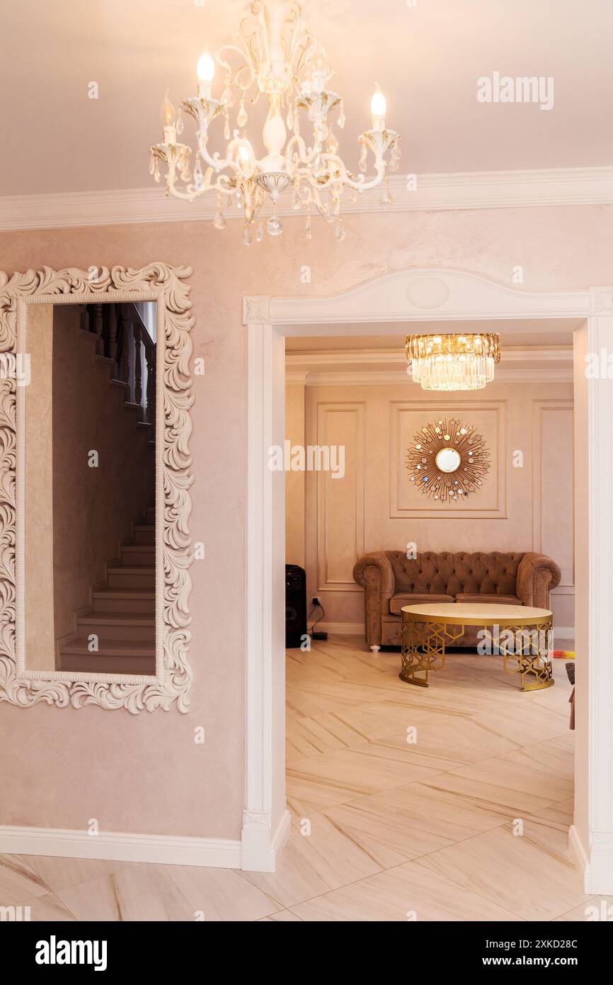 Un élégant couloir avec un lustre et un miroir orné sur un mur rose clair, menant à un salon élégant avec un canapé et un miroir décoratif. Banque D'Images