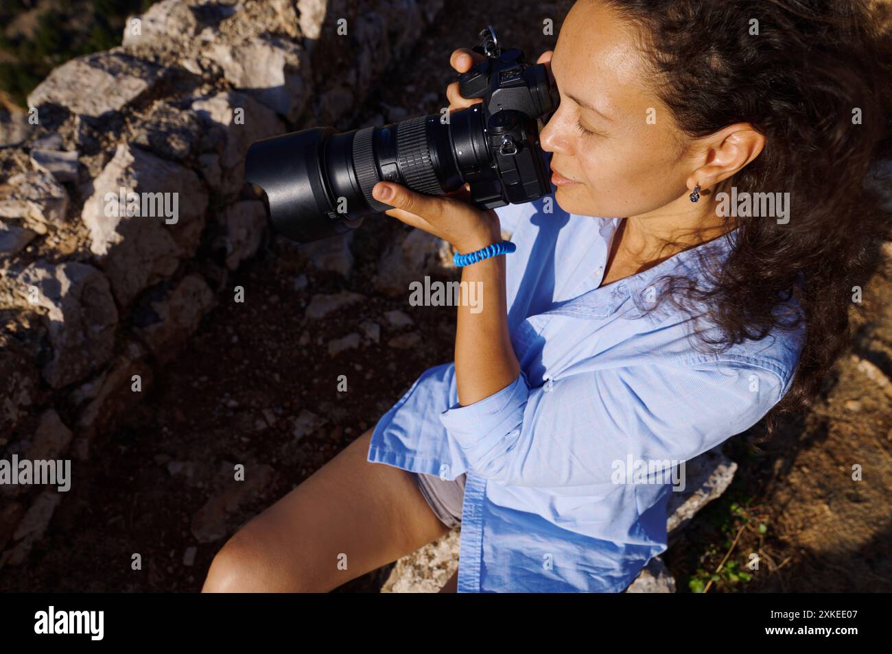 Une femme photographe capture de superbes moments en extérieur avec son appareil photo reflex numérique assis sur une montagne rocheuse. Elle est concentrée et immergée dans sa photo Banque D'Images