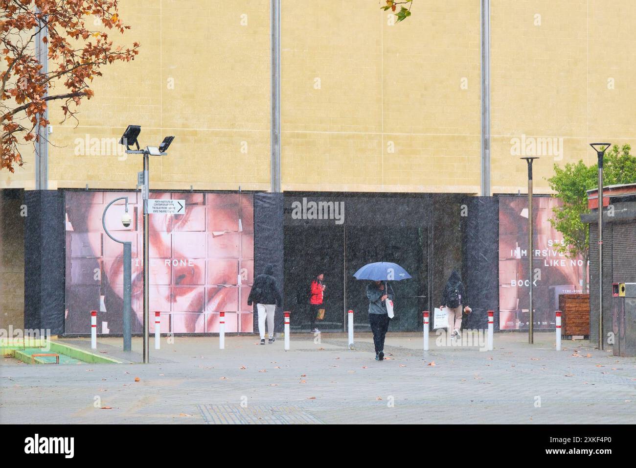 Des gens marchant sous la pluie en hiver devant la galerie d'art, l'un avec un parapluie, au Perth Cultural Centre à Northbridge, Perth, Australie occidentale. Banque D'Images