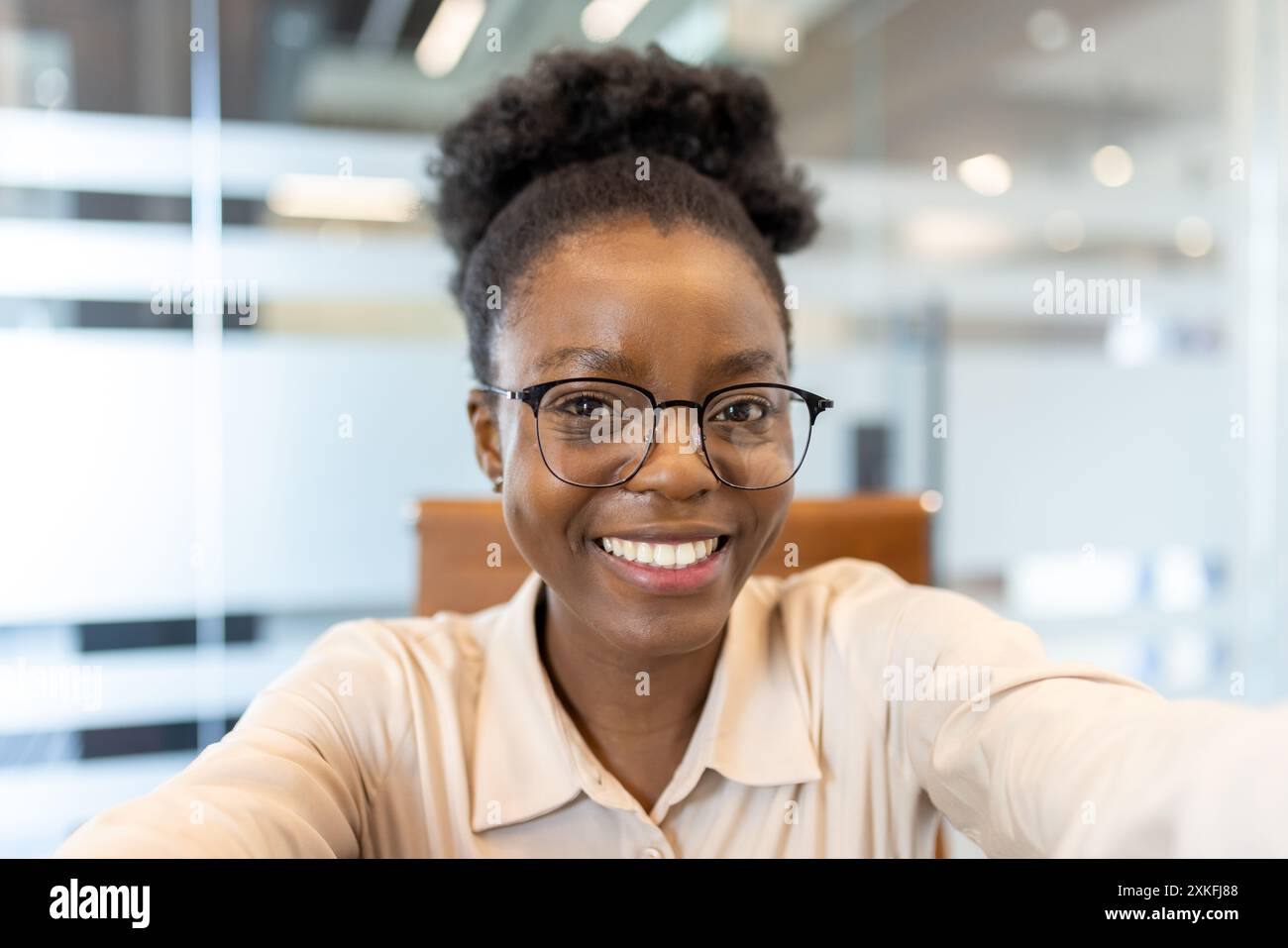 Femme souriante portant des lunettes prenant selfie dans un bureau moderne pendant un appel vidéo. Capture les émotions positives, le cadre professionnel et la communication virtuelle. Banque D'Images