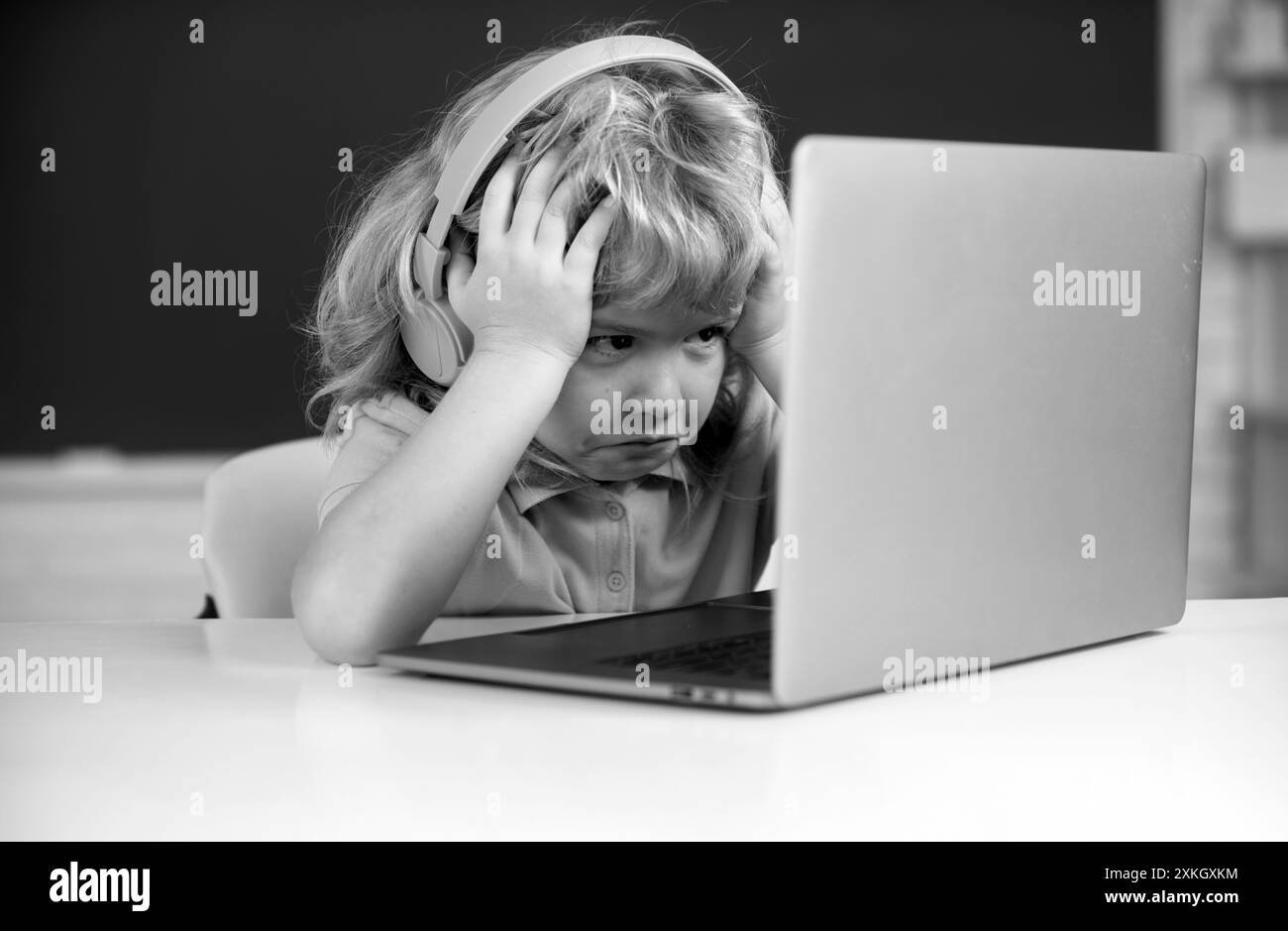 Un garçon triste en colère dans un casque s'assoit au bureau, étudie en ligne sur un ordinateur portable à l'école, klever enfant porte des écouteurs dans l'apprentissage d'un ordinateur portable en utilisant des leçons d'Internet. Banque D'Images