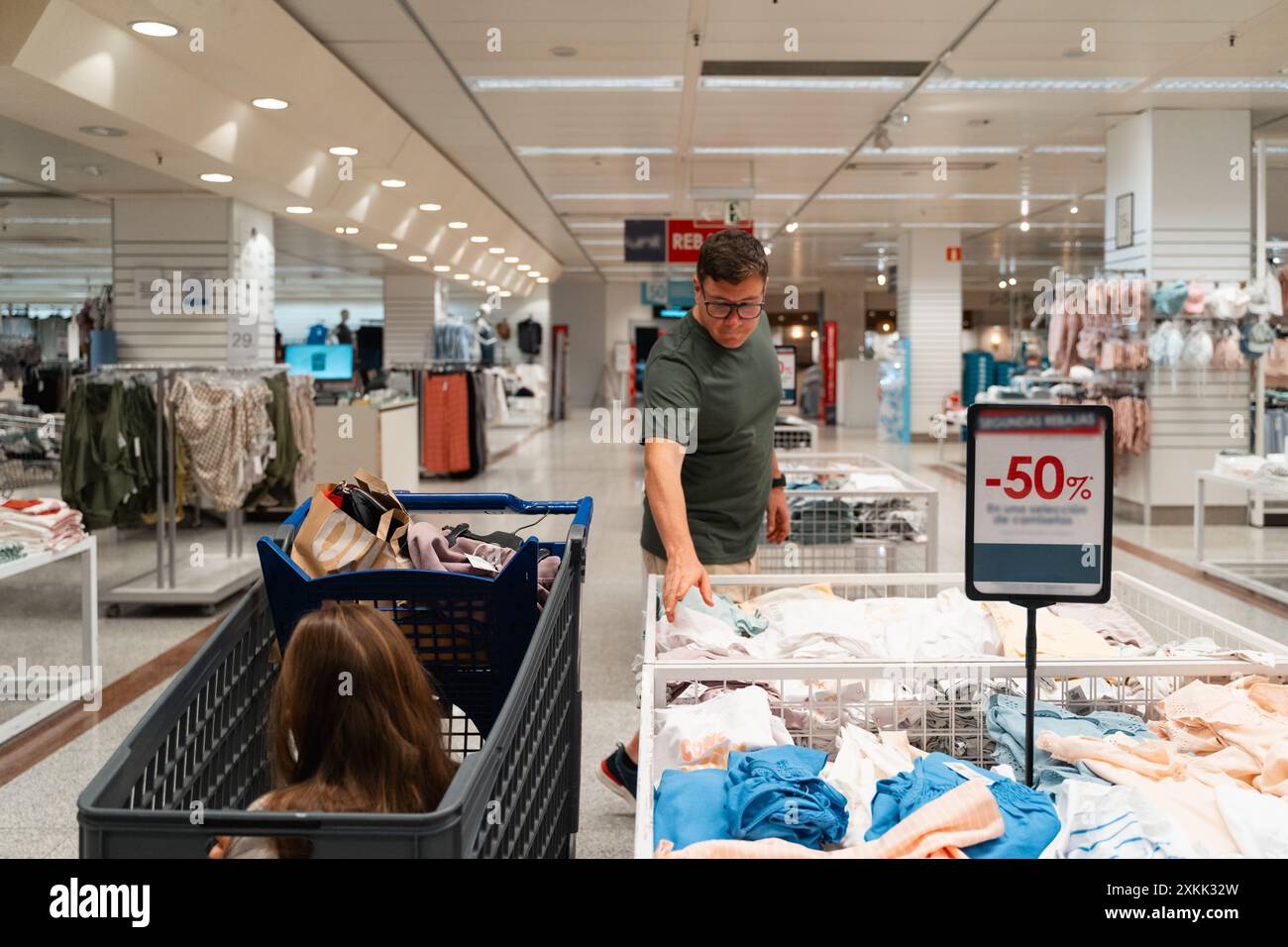 Un homme magasinant des vêtements dans un magasin pendant que son enfant est assis dans le chariot. Il examine un rack de vêtements à prix réduit. Consommateur masculin, chasseur de bonnes affaires Banque D'Images