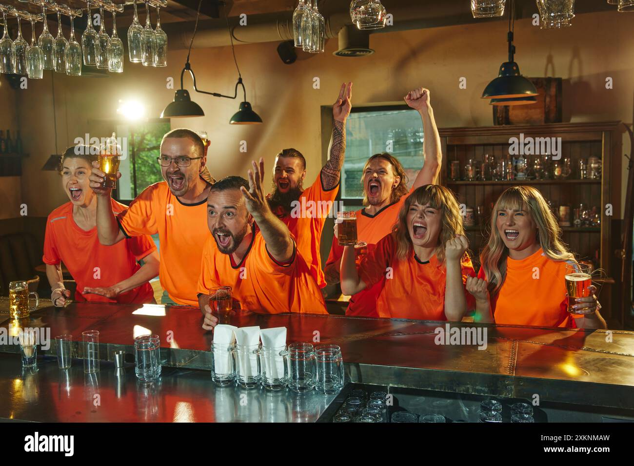 Les fans enthousiastes vêtus de chemises orange s'assoient au bar avec des verres de bière, regardant des événements sportifs et montrant des expressions de célébration. Banque D'Images